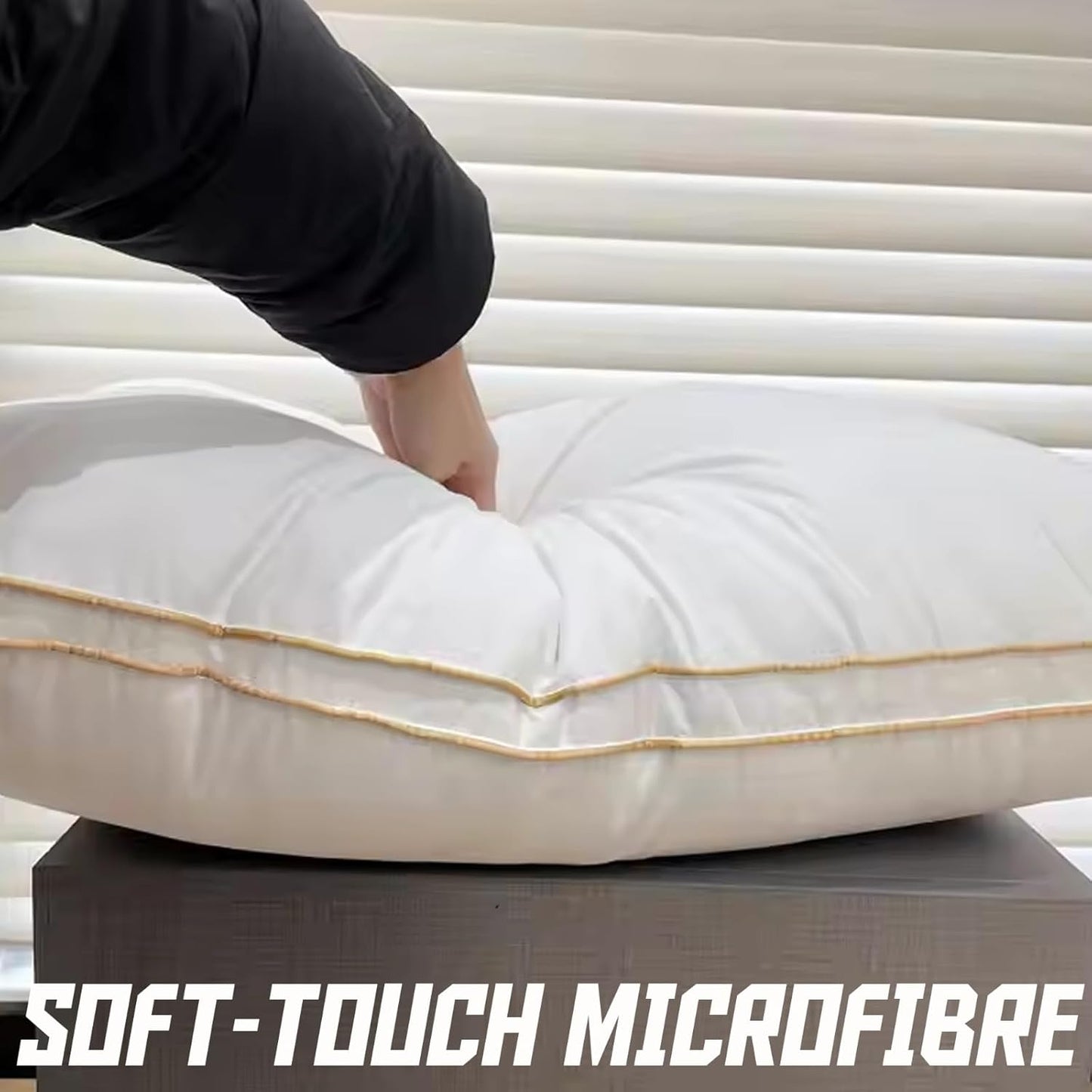White pillow with gold trim being held by a hand against white blinds, with 'Soft-Touch Microfibre' text at the bottom.