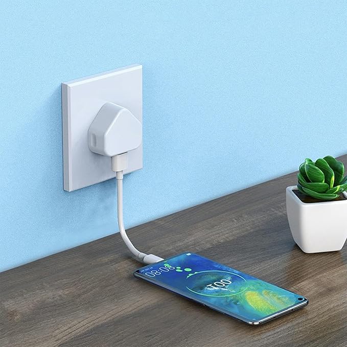 Phone being charged via a cable connected to a wall outlet on a wooden surface with a plant in the background.