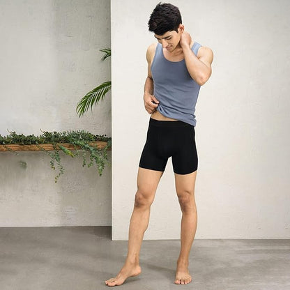 Man wearing a gray tank top and black boxers standing against a plain wall with a plant.