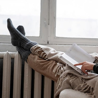 Person relaxing with feet up on a radiator, reading a book, wearing a beige blanket and dark socks.