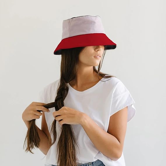 Woman wearing a white and red bucket hat, braiding her hair against a plain background