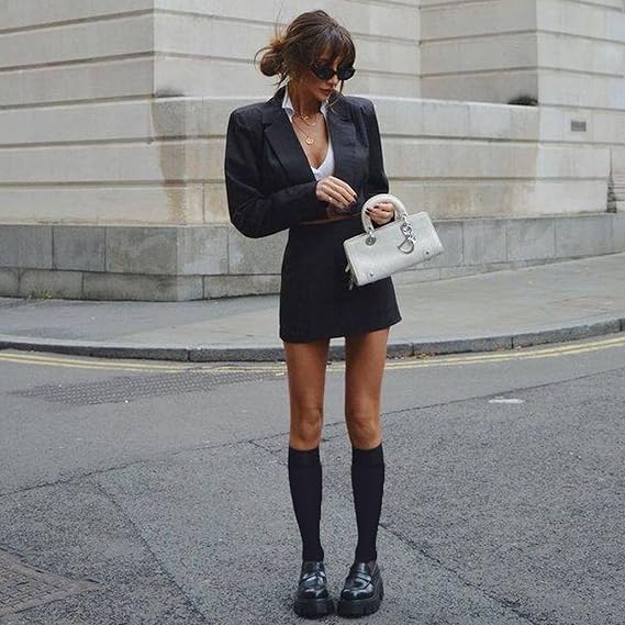 Woman in a black blazer and skirt with knee-high socks and black shoes, holding a white handbag on a city street.