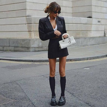 Woman in a black blazer and skirt with knee-high socks and black shoes, holding a white handbag on a city street.